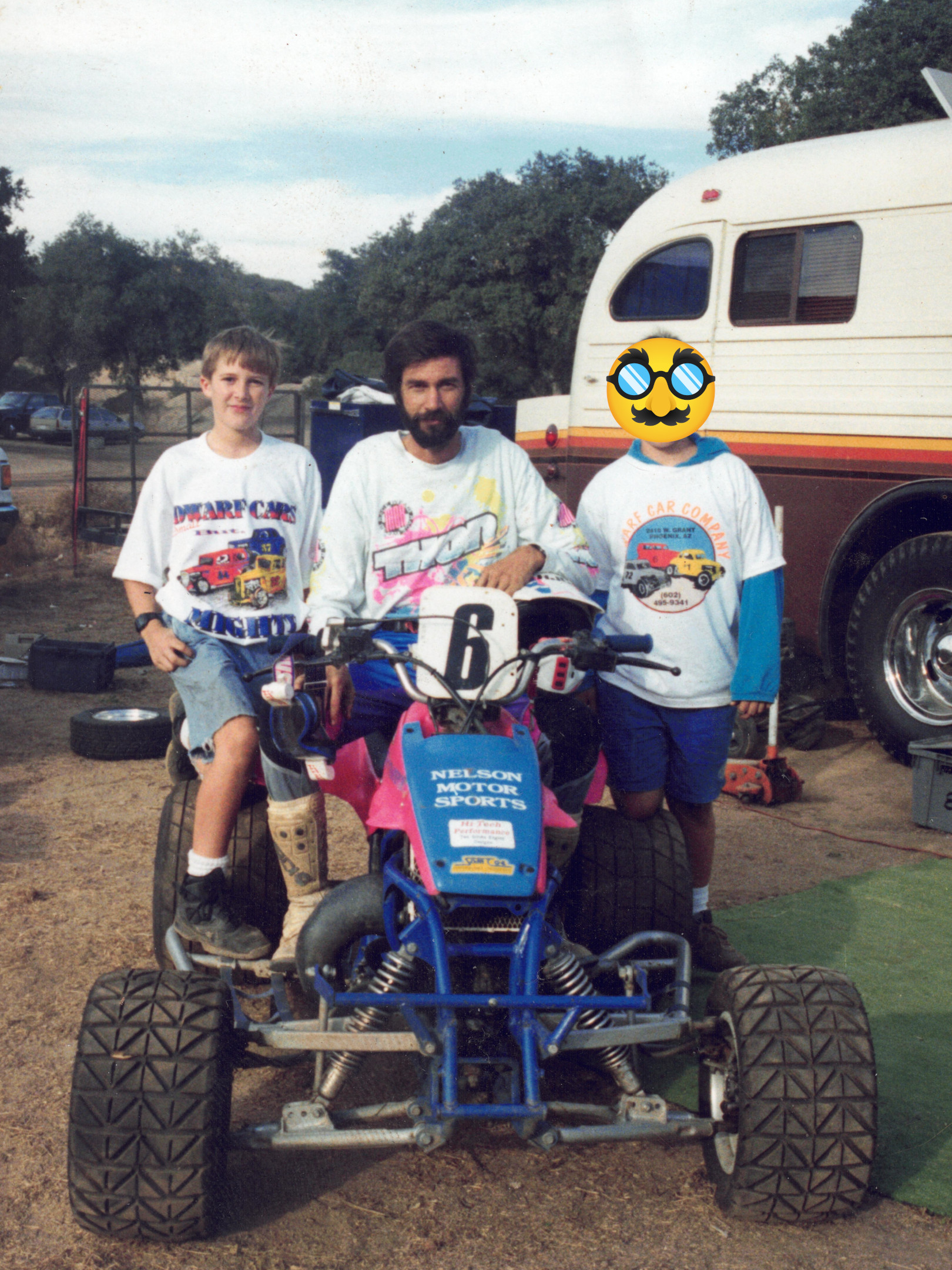 Man with dark hair and beard sitting on a racing quad with a young boy of maybe 12 years old standing next to him. Another boy is on the other side but obscured by a disguise emoji